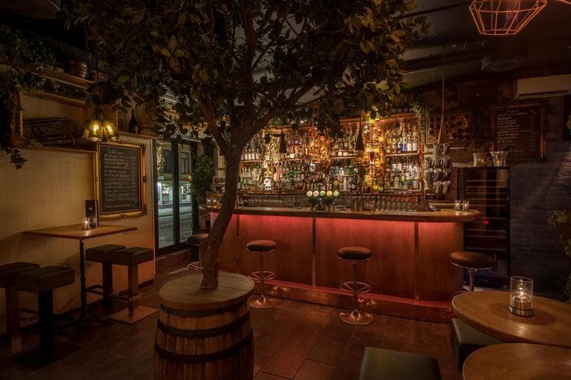 Barinteriør med barbenk, barstoler, små bord og stoler, og et tre som står i en tønne midt i rommet.