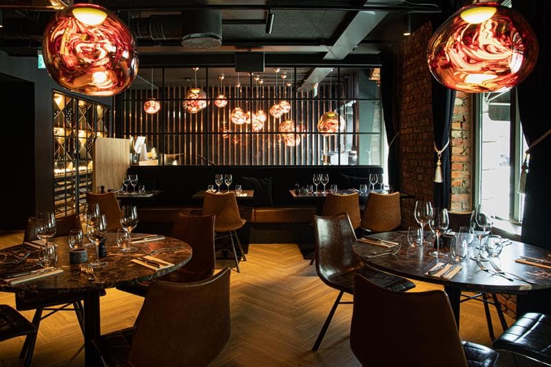 Restaurantinteriør med runde, brune bord, brune stoler og sitteseksjon mot veggen.