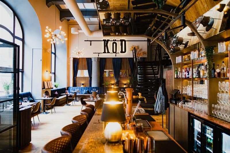 Restaurantinteriør med bardisk, barstoler, hyller med glass og vinflasker, blå sitteseksjon langs veggen og høyt under taket